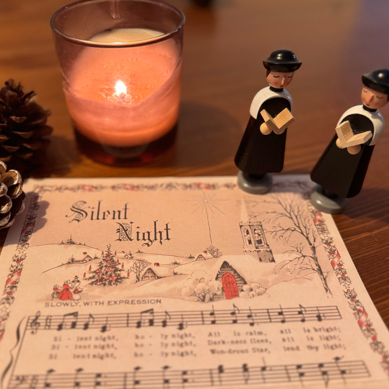 Two small figurines standing on a sheet of 'Silent Night' music with a lit candle in the background.