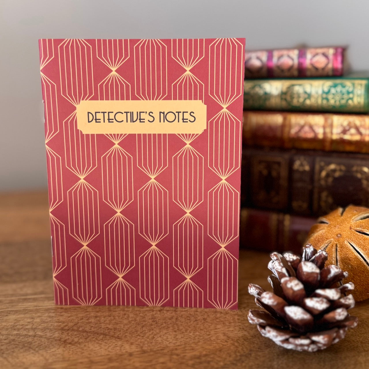 Notebook titled 'Detective's Notes' with Art Deco pattern on a wooden surface with books and a pine cone.