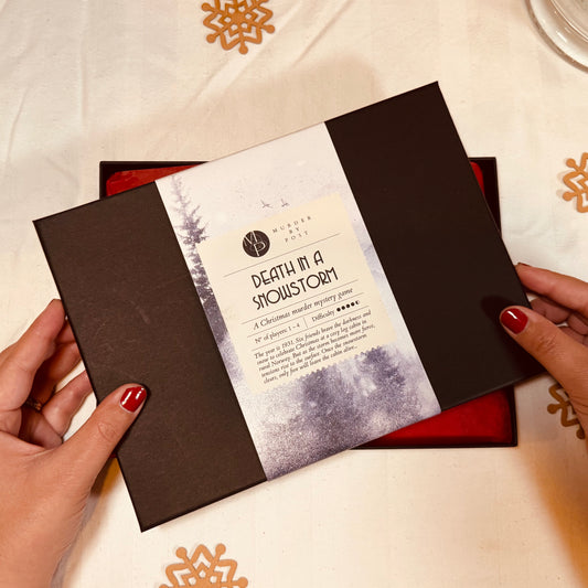 A person's hands with red nail polish hold a black lid of the murder mystery advent calendar 'Death in a Snowstorm'. The book has a white paper label wrapped around the center with black text and a snowy forest illustration.