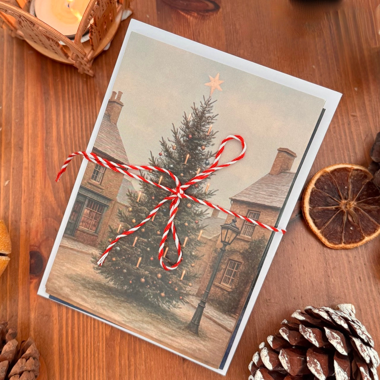 Vintage-style Christmas card set, the top one with a tree and house, tied with red string on a wooden surface with pine cones and an orange.