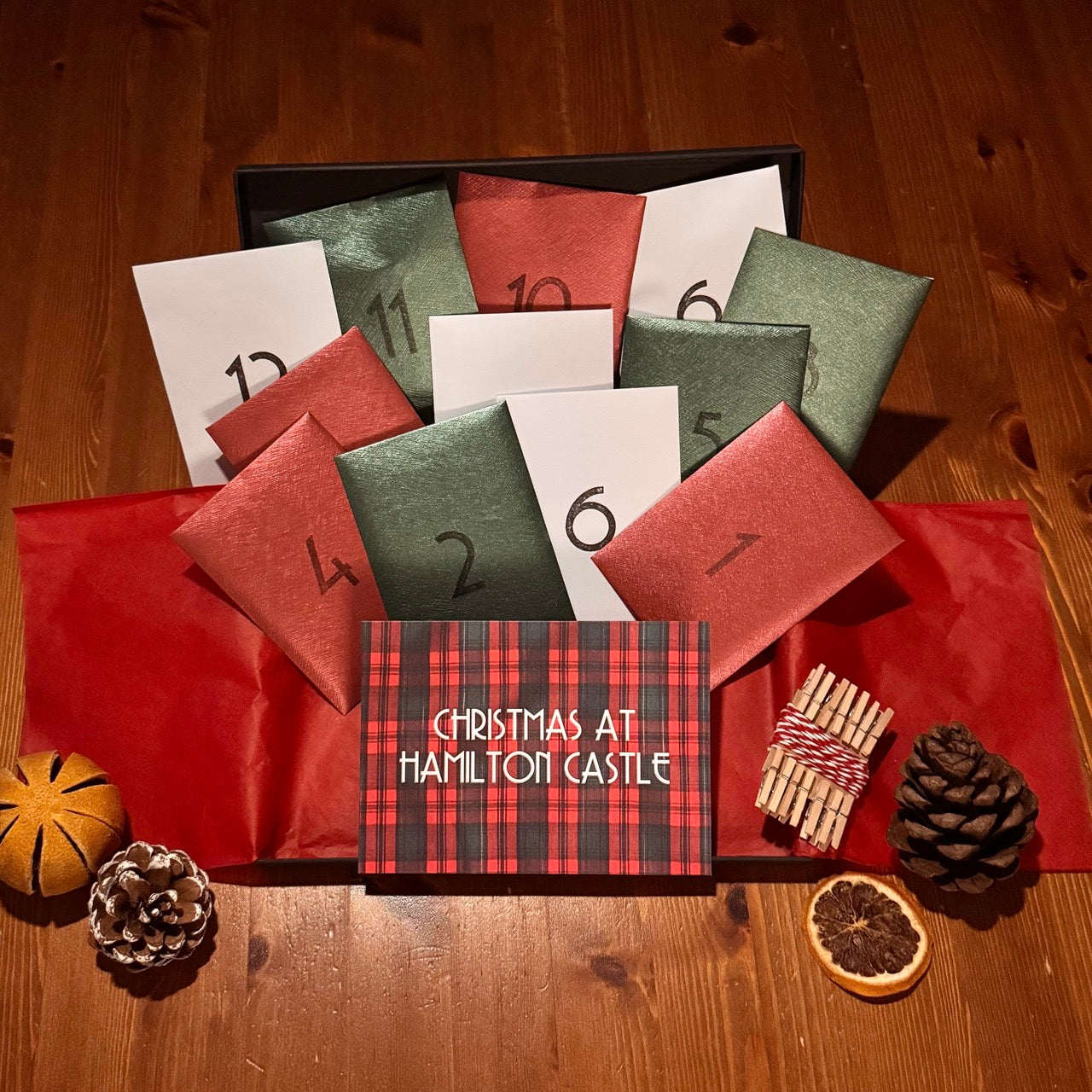An open Murder Mystery Advent Calendar box arranged on a wooden tabletop, with 12 numbered envelopes popping out of the box (green, red and white). A card at the front reads 'Christmas at Hamilton Castle.' At the front of the box are a few festive decorative items including pinecones, a dried orange and dried orange slice. 