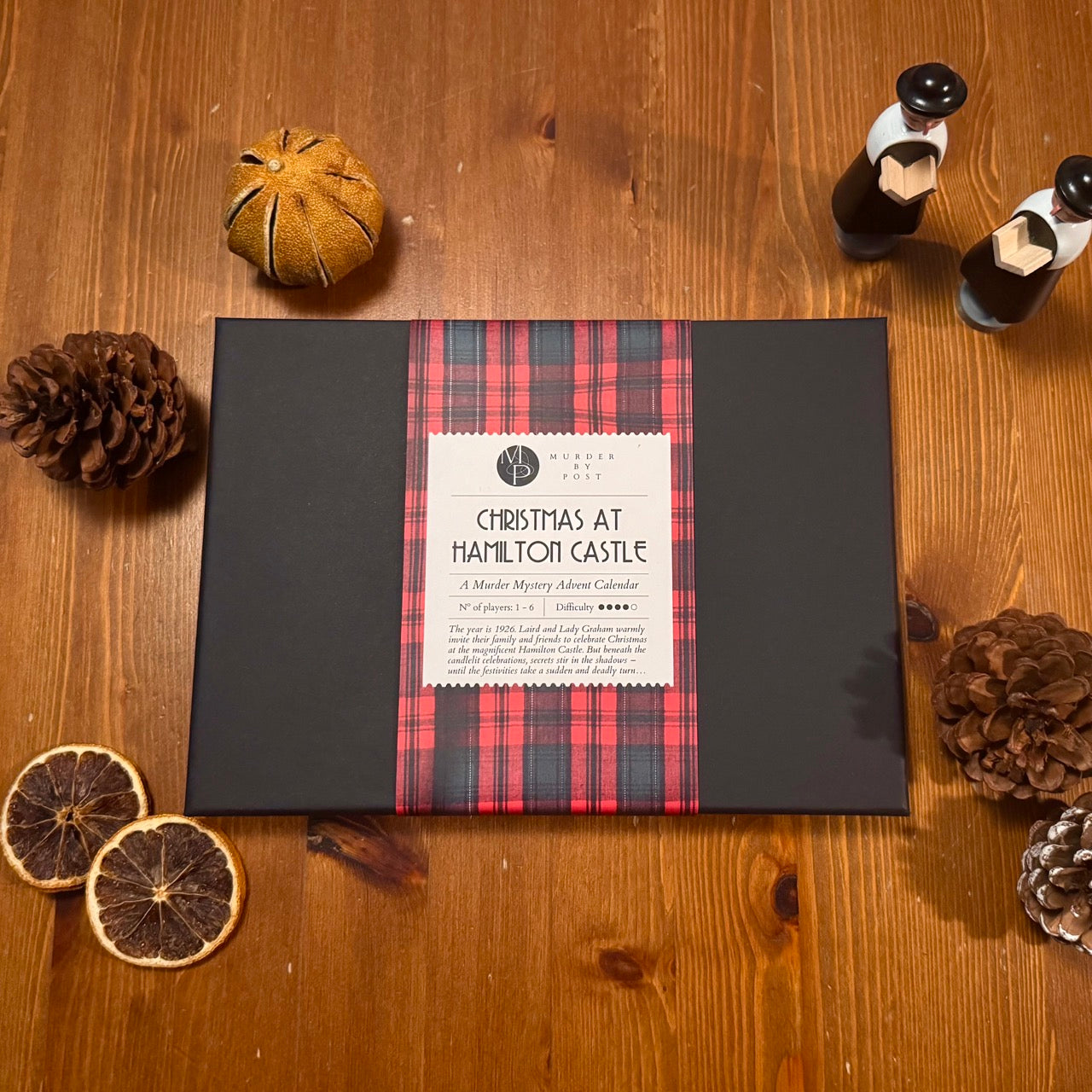 A black game box with a red and green tartan bellyband and a white label reading 'Christmas at Hamilton Castle,' styled on a wooden surface decorated with pinecones, dried orange slices, and wooden Christmas ornaments.