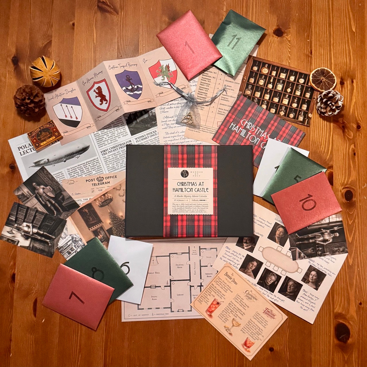 The central black game box of the Murder Mystery Advent Calendar 'Christmas at Hamilton Castle' features a red and green tartan ribbon with a white label. The box is surrounded by the contents of the Advent Calendar spread across a table, including shiny envelopes, a table plan, a newspaper, a pendant in a small pouch, a cocktail recipe, old photographs of suits of armour, and several other items. 