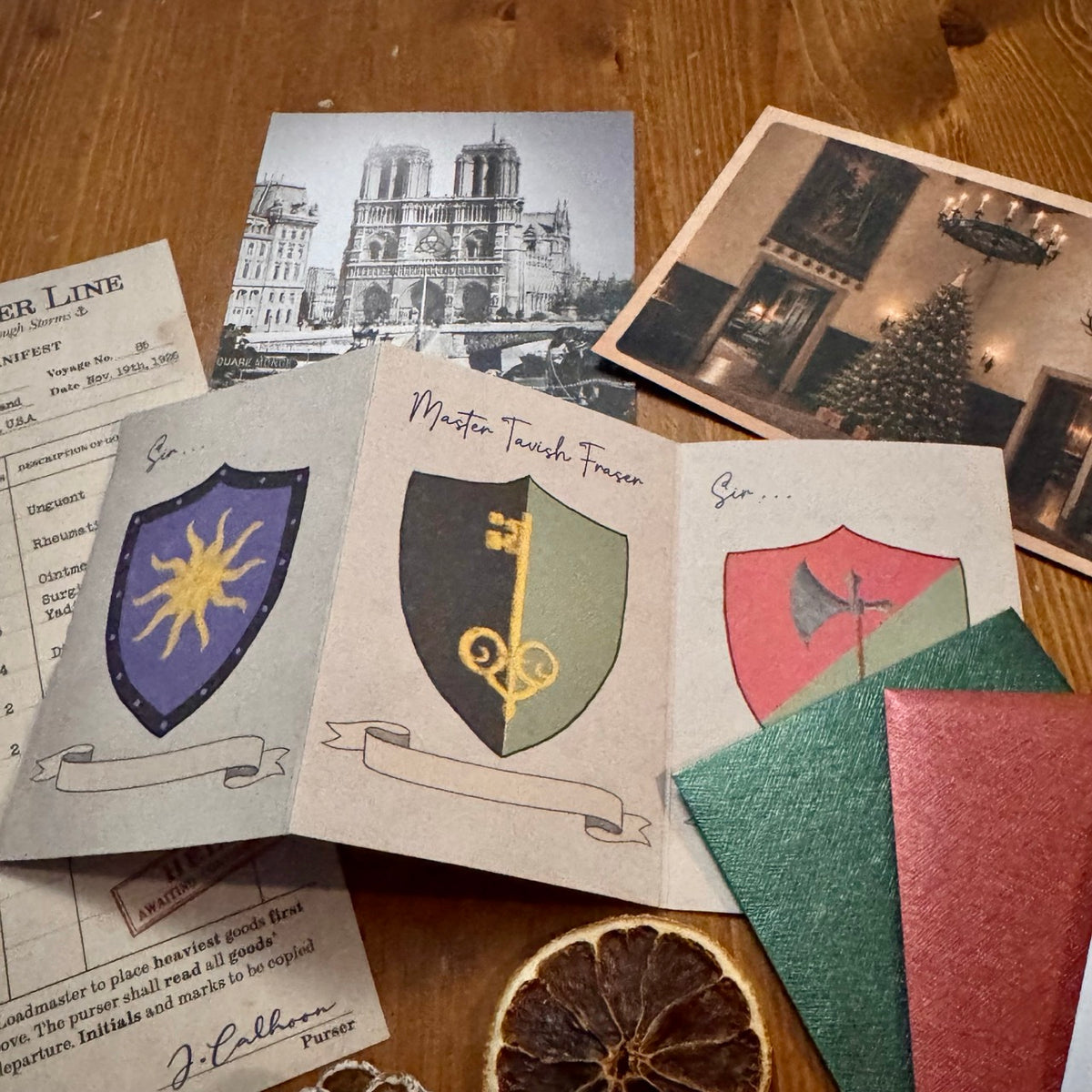 A styled layout of vintage-themed cards and papers on a wooden table, all parts of the Advent Calendar. One concertina fold paper feature medieval-style heraldic shields, each with a gold key, a sun, and an axe, alongside a black-and-white photo of Notre Dame Cathedral and a Christmas scene.