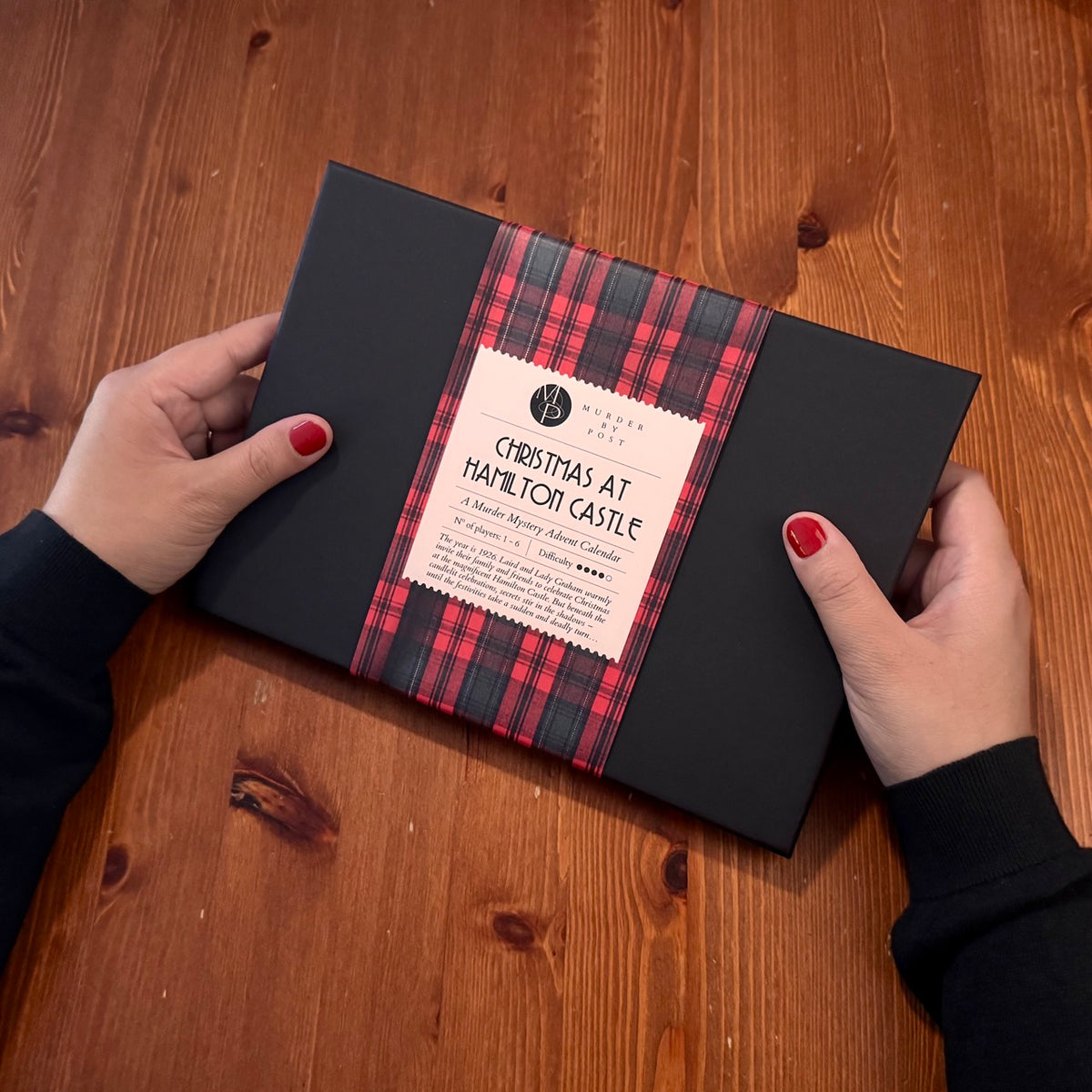 A black rectangular game box wrapped with a red and green tartan bellyband and a cream label reading 'Christmas at Hamilton Castle,' held by two hands over a wooden tabletop.