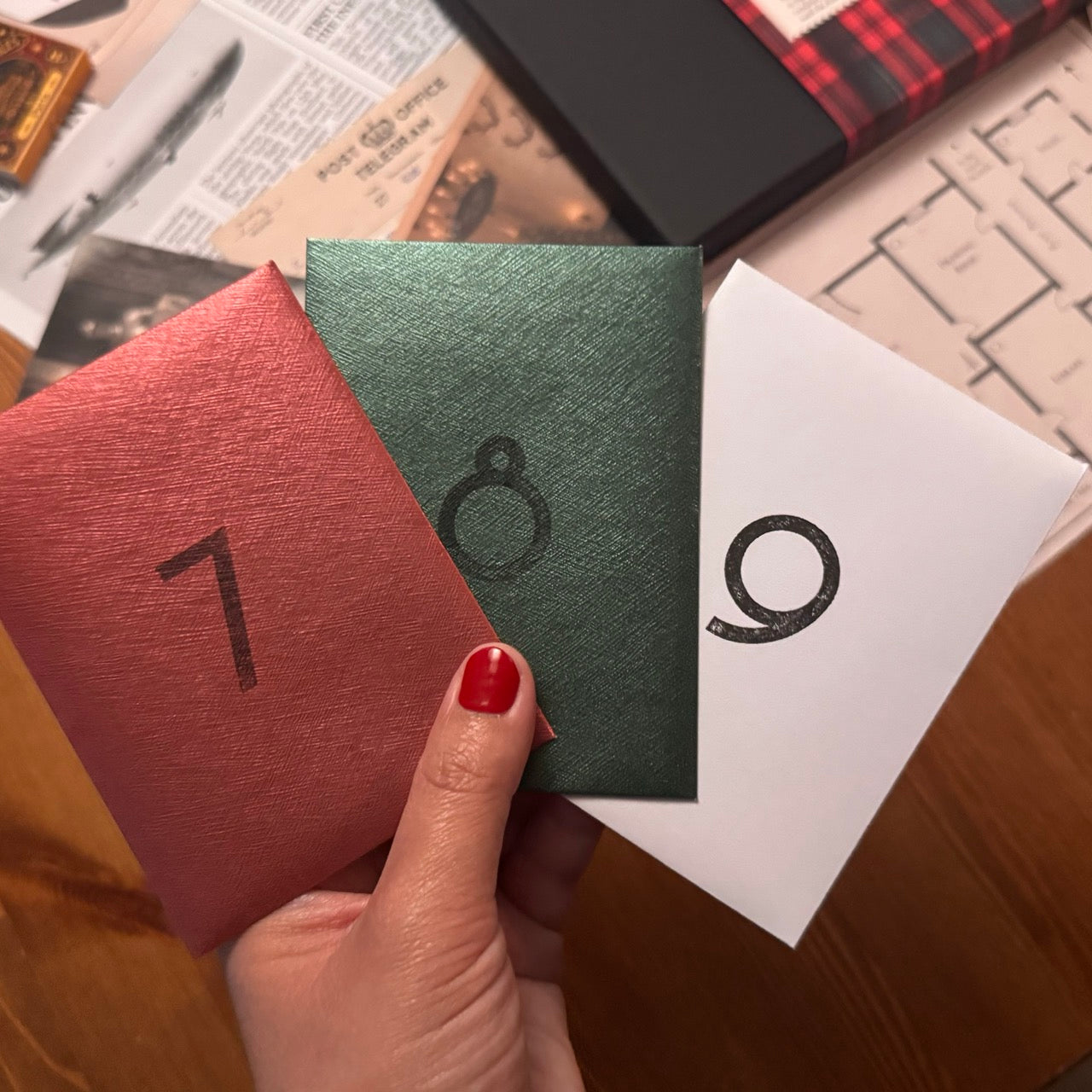 A hand with red nail polish holds three textured paper envelopes in red, green, and white, each printed with a black number. The background is a wooden table strewn with papers and notes. 