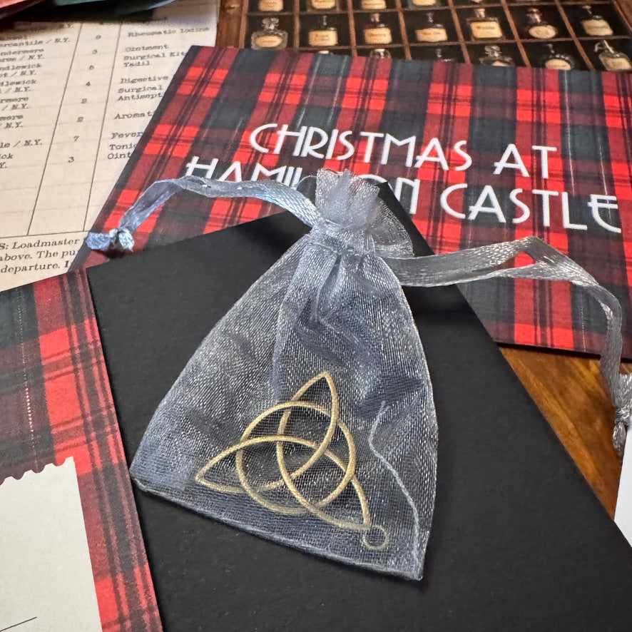 A small sheer gray organza drawstring pouch with a gold Celtic triquetra symbol, placed on a black card over a red plaid and white printed insert.