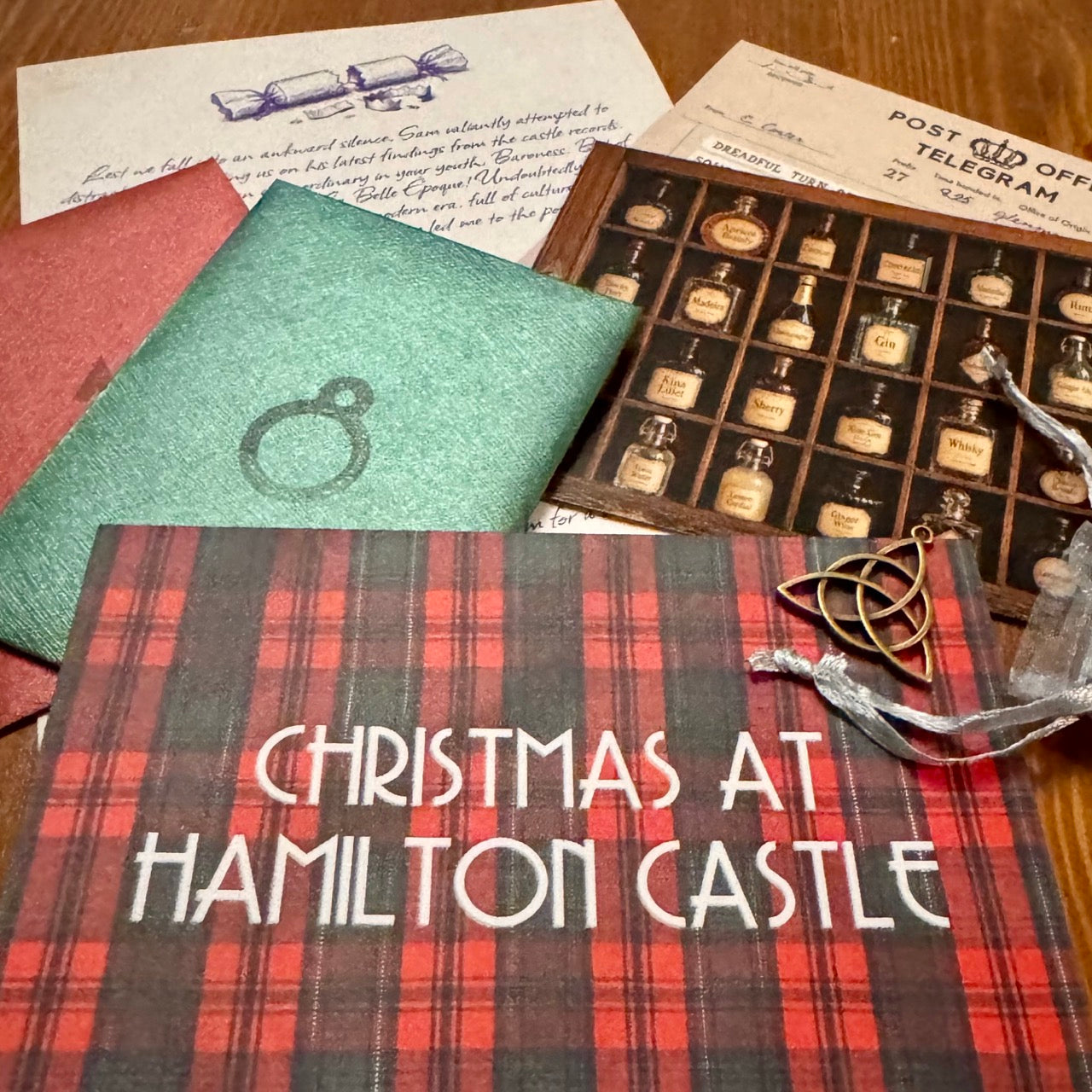 A themed flat-lay of murder mystery advent calendar game pieces, featuring a red and green tartan card with white text, a small brass pendant in the shape of a Triquetra, a photograph of a wooden cabinet of alcohol bottles, shiny green and red numbered envelopes.