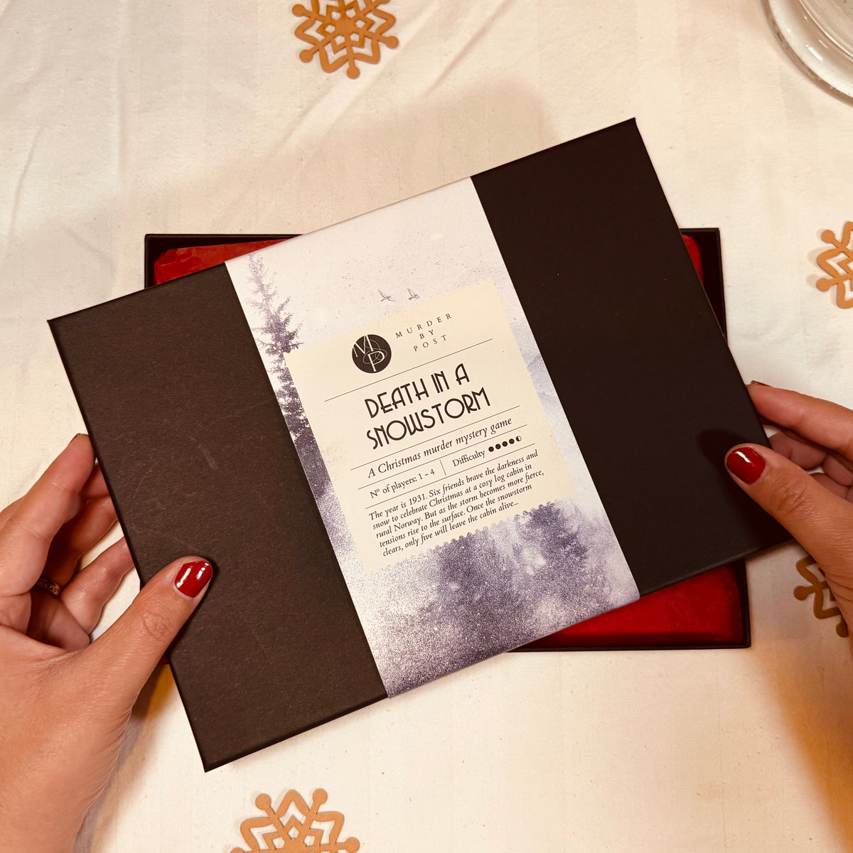 A person's hands with red nail polish hold a black lid of the murder mystery advent calendar  'Death in a Snowstorm'. The book has a white paper label wrapped around the center with black text and a snowy forest illustration.