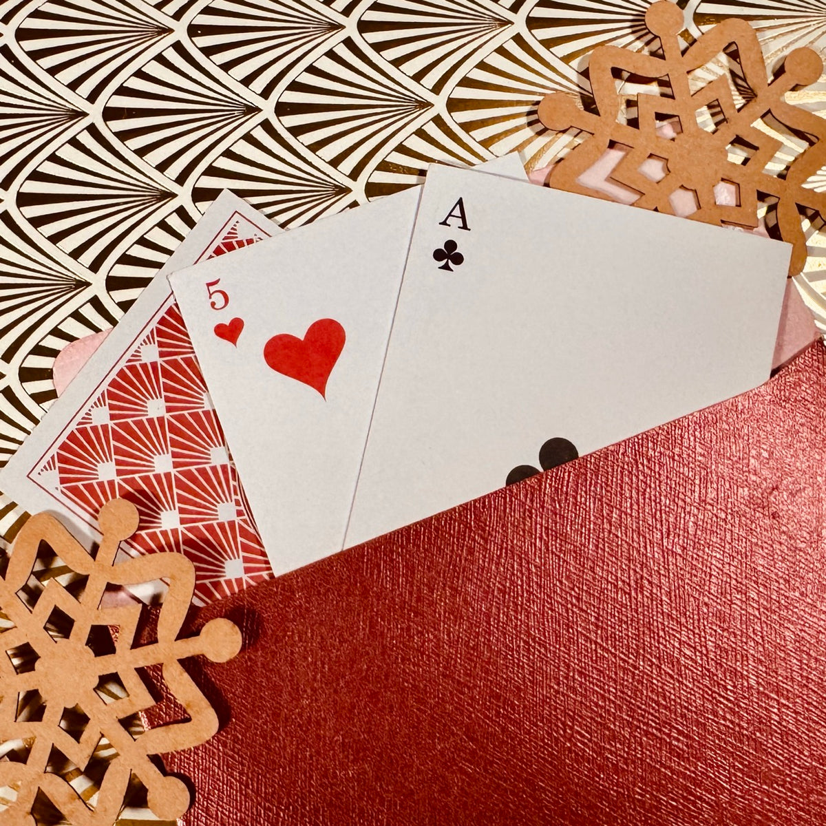 Three playing cards fanned out from a textured red envelope on a patterned gold and black background with decorative snowflakes.