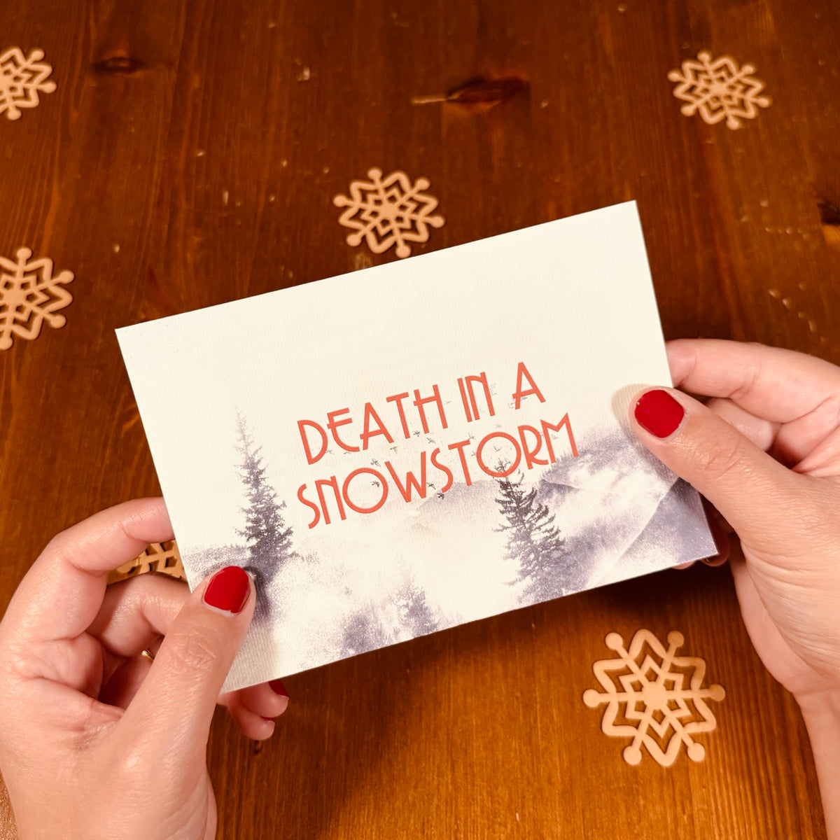 A person holds a card with the text 'DEATH IN A SNOWSTORM' in coral-red lettering over a snowy forest illustration. The card is photographed over a warm brown wooden surface with laser-cut snowflake decorations.