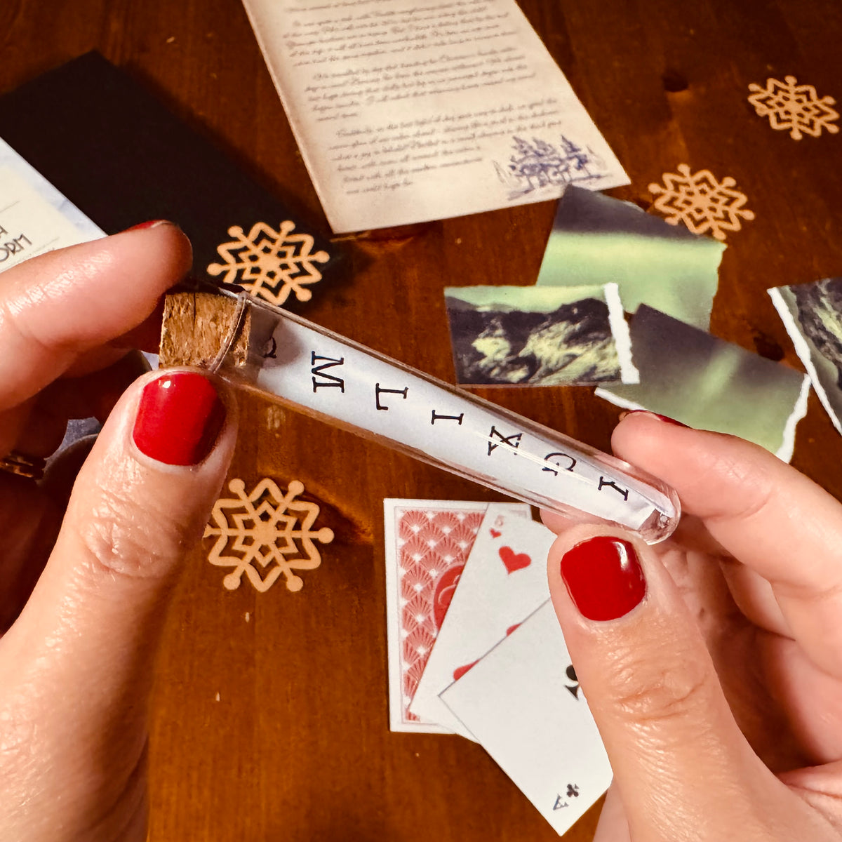 A close-up of hands with red nail polish holding a clear glass vial with a cork stopper containing a piece of paper with text. The vial is shown over a wooden tabletop with scattered decorative elements including wooden snowflakes, playing cards, and paper scraps.